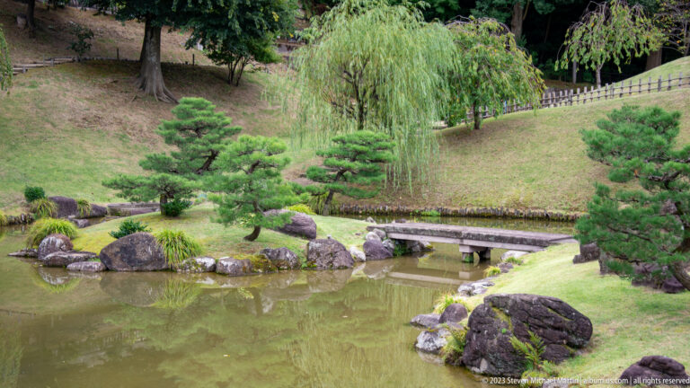 Low stone crossing over a garden pond with clipped pines, boulders, shrubs, and a weeping tree.
