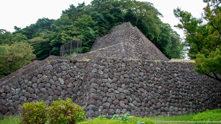 Tall angled stone retaining walls built from irregular dark blocks beside a narrow path and dense greenery.