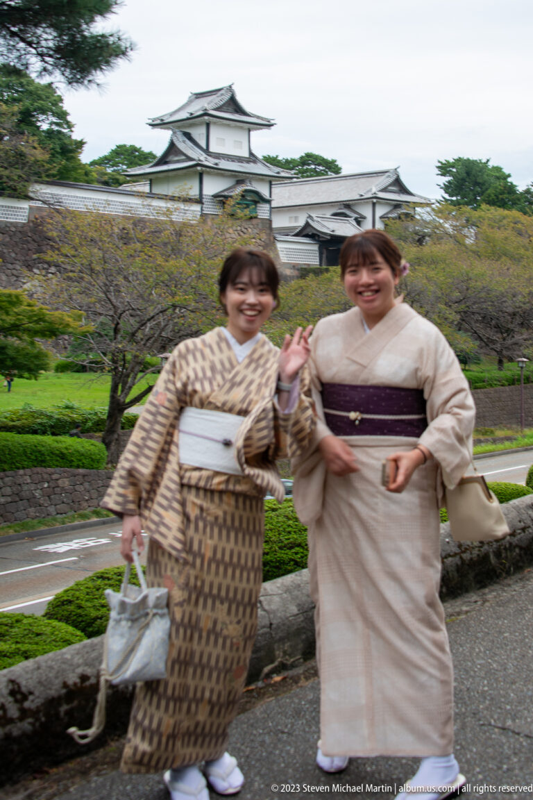 Two smiling women in patterned kimono pose before white castle buildings, tiled roofs, stone walls, and landscaped grounds.