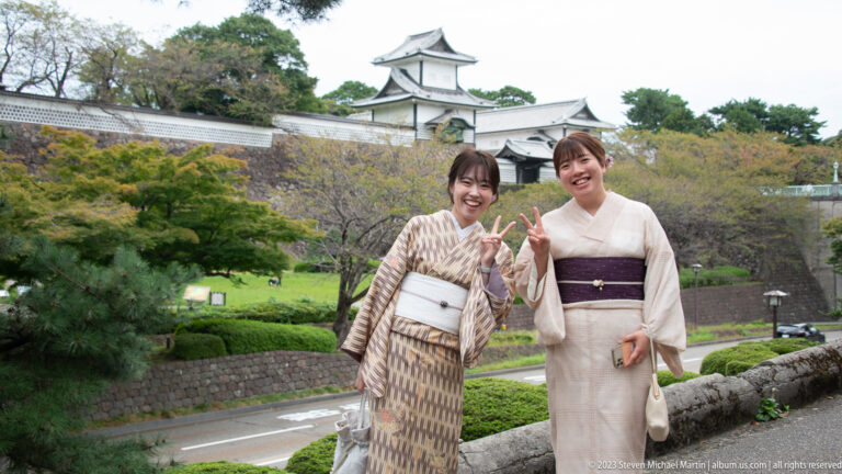 Two women in patterned kimono smile and make peace signs with a castle turret and stone walls behind them.