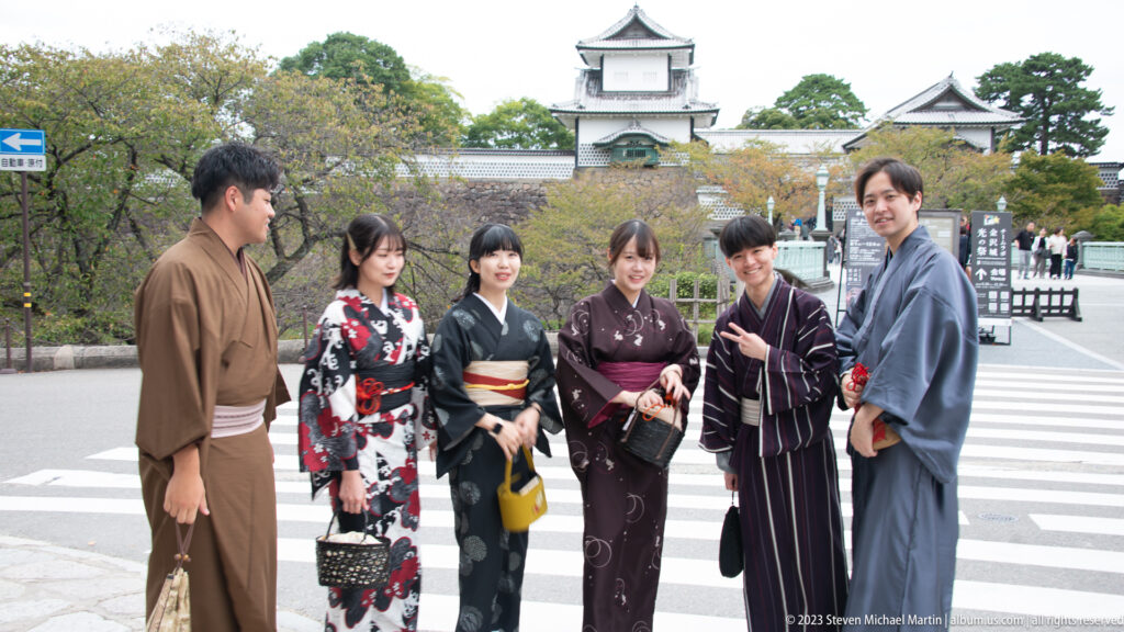 Six visitors in varied kimono pose near a castle approach beneath a white turret and stone rampart.