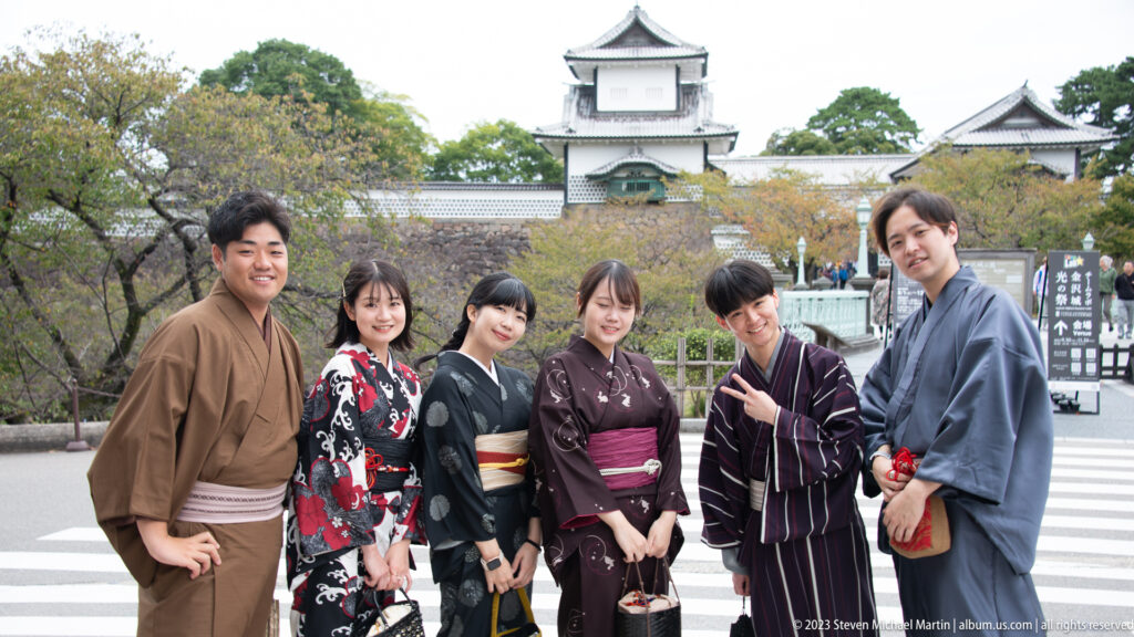 Close group portrait of six visitors in patterned kimono with stone walls and a white castle turret behind them.