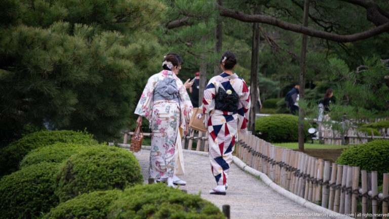 Two women seen from behind walking along a garden path in colorful patterned kimono among pines and clipped shrubs.