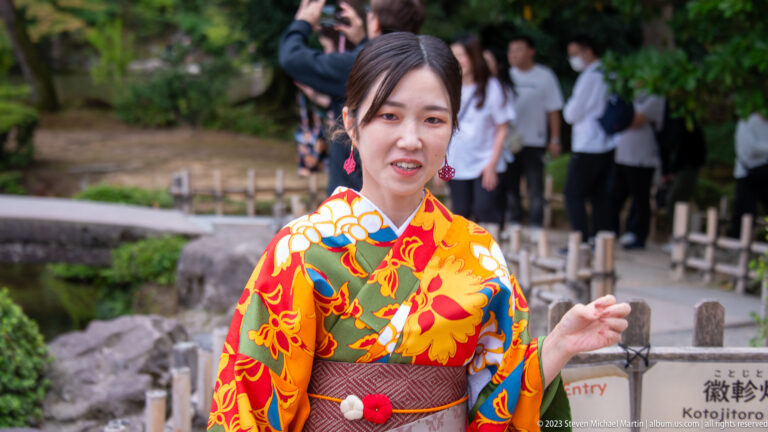 Woman in a bright orange-red floral kimono standing beside a garden path with visitors and signage behind her.
