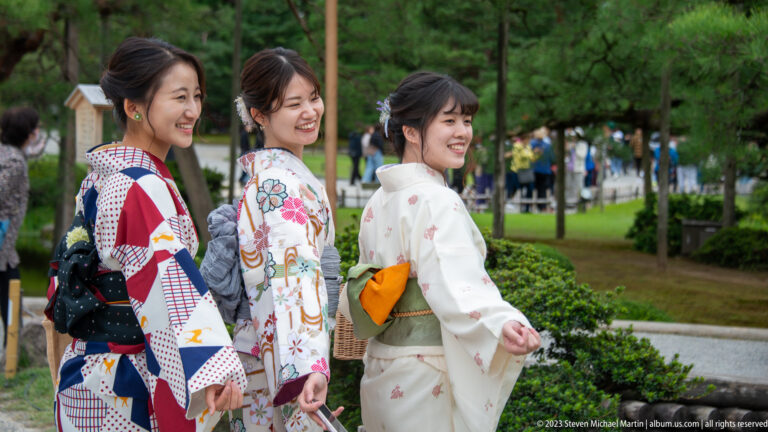Three smiling women in patterned kimono look back toward the camera on a path through a landscaped Japanese garden.