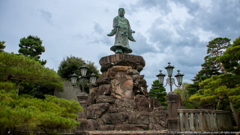 Bronze robed figure stands on a tall rough-stone pedestal among pines, lamps, and a stone railing.