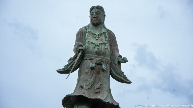Close view of a green-patinated bronze robed figure with both hands extended against a cloudy sky.