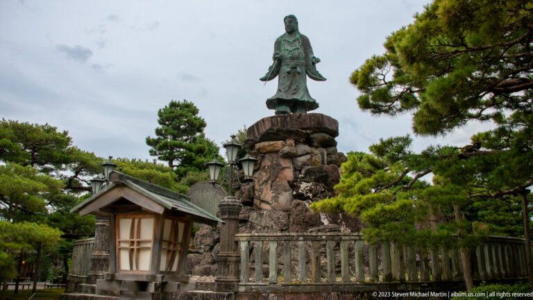 Bronze statue on a rugged stone pedestal framed by shaped pines, a lantern structure, and a stone railing.