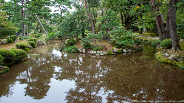 Reflective pond edged with mossy rocks, trimmed shrubs, and mature trees in a shaded Japanese garden.