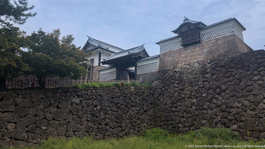 White castle turrets and tiled roofs rise above a high sloping wall of dark irregular stone.