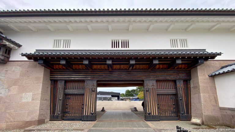 Large dark wooden gate beneath a tiled canopy in a white castle wall with small barred openings.
