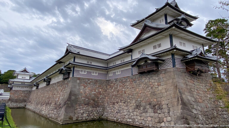White multi-level castle turrets with tiled roofs stand above a sloped stone rampart beside a moat.