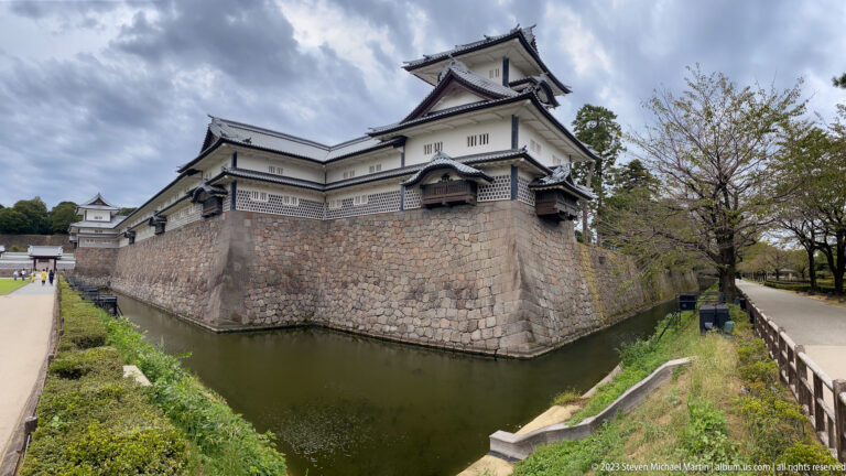 White castle turrets rise above a corner stone wall and moat, with a pedestrian path at right.