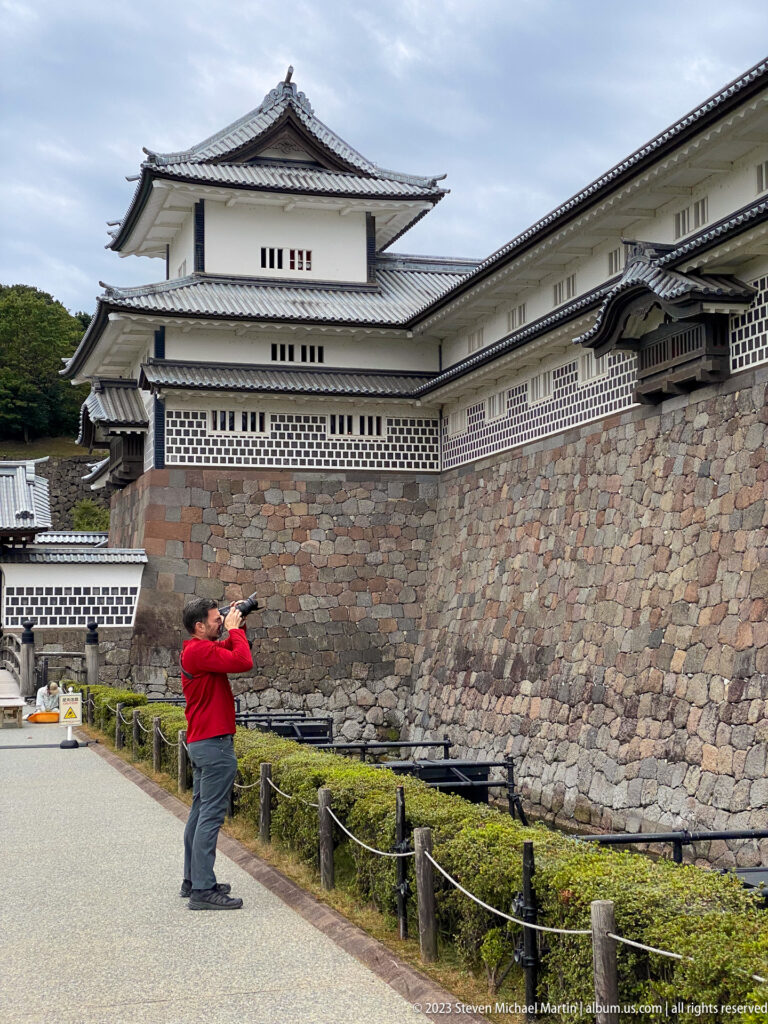 Visitor in a red shirt photographs white castle buildings above a massive sloped stone wall.