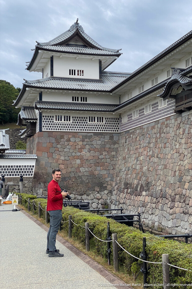 Visitor in a red shirt stands before a high stone castle wall and white tiled-roof buildings.