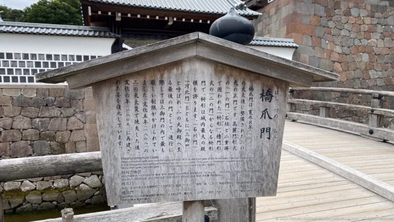 Bilingual interpretive marker labeled Hashizume-mon Gate stands beside a bridge and stone castle walls.