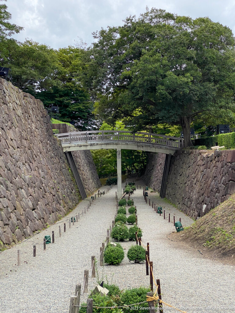 Long gravel passage with clipped shrubs descends between high stone walls beneath a pedestrian bridge.