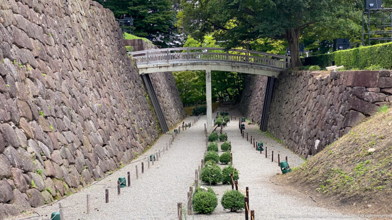 Gravel corridor and clipped shrubs lead between steep stone ramparts toward a bridge overhead.