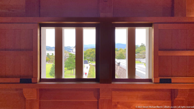 Narrow barred windows in a warm wooden castle interior overlook lawns, roofs, and distant hills.
