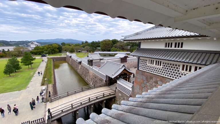 Elevated castle view over a moat, bridge, white walls, lawns, and distant wooded hills.