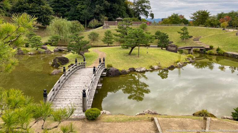Arched wooden bridge crosses a garden pond surrounded by shaped pines, rocks, and sloping lawns.