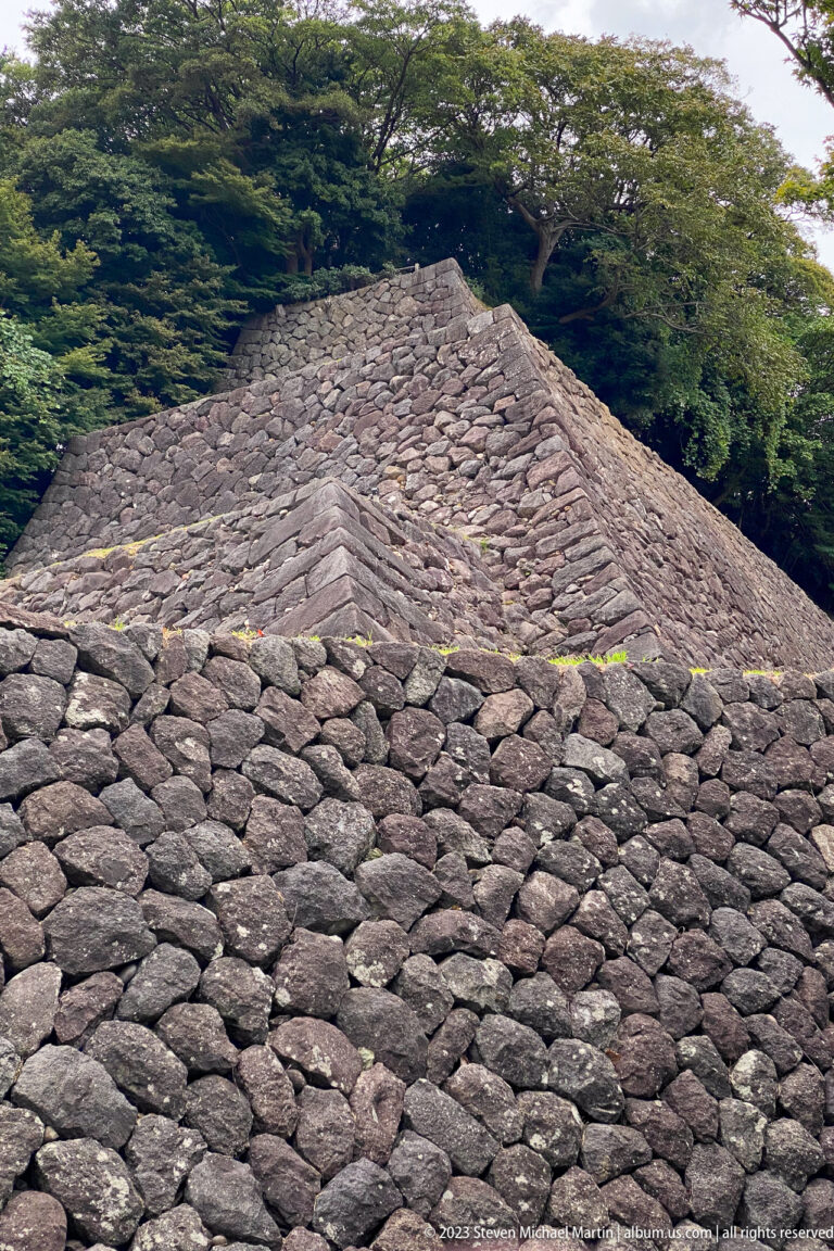 Steep castle wall of tightly packed irregular stones rises to a vegetated crest.