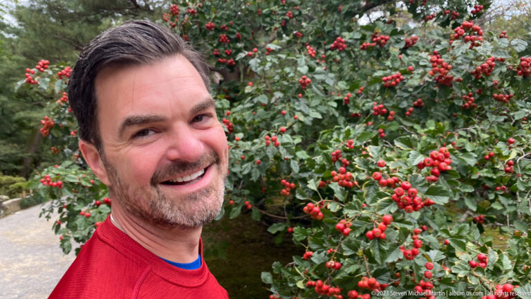 Smiling man in a red shirt poses beside dense green foliage covered with bright red berries.