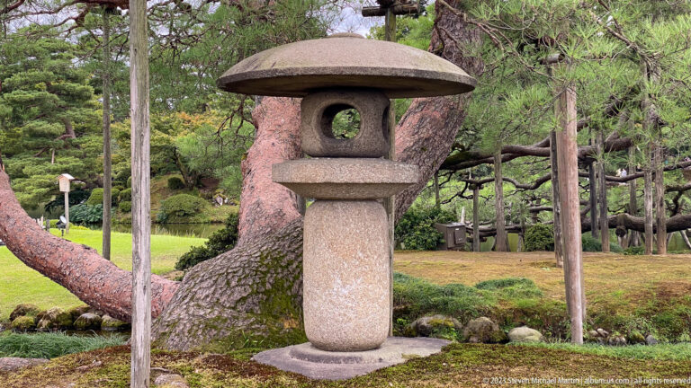 Traditional stone lantern with broad cap stands on moss beneath mature pines and wooden support poles.