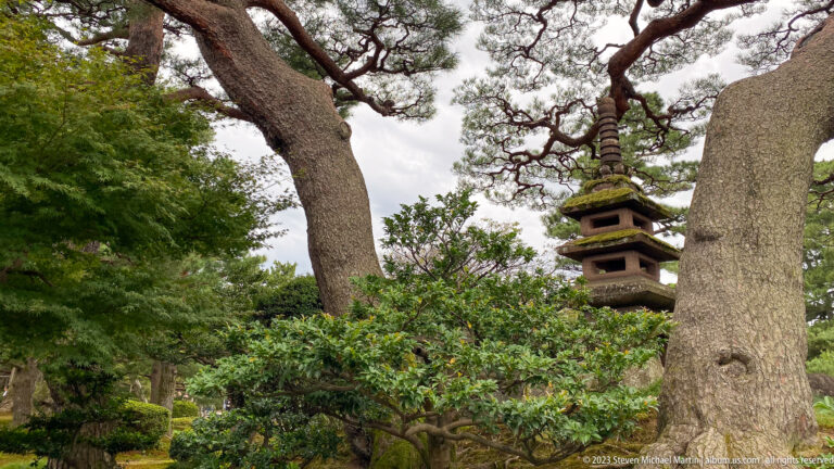Multi-tiered stone pagoda rises behind mature tree trunks and dense foliage in a Japanese garden.