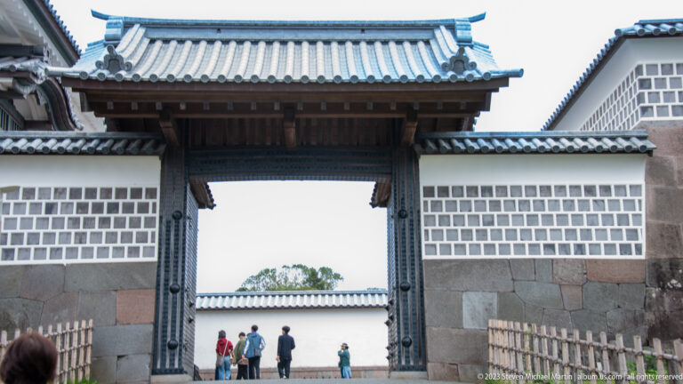 Tall timber gate in white plaster castle walls, with visitors walking through the opening.