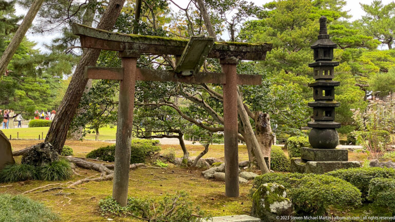 Weathered torii and tall stone pagoda stand together among moss, shrubs, and mature garden trees.