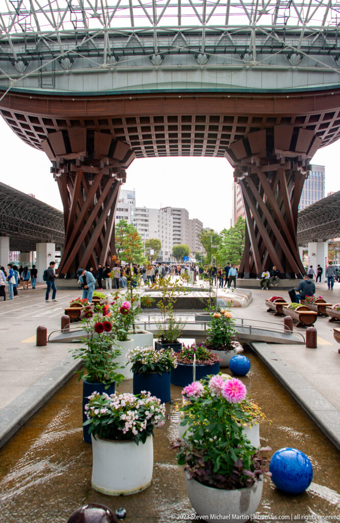Tsuzumi-mon Gate at Kanazawa Station with massive wooden columns, glass canopy, reflecting pools, flowers, and pedestrians.