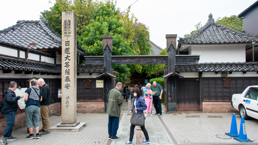 Visitors standing at Myōryū-ji’s timber entrance beside a tall inscribed stone pillar and tiled walls.