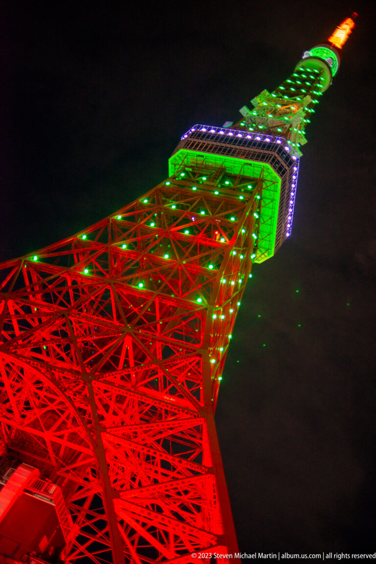 Extreme upward view along Tokyo Tower’s red lattice to its green upper section and orange tip.