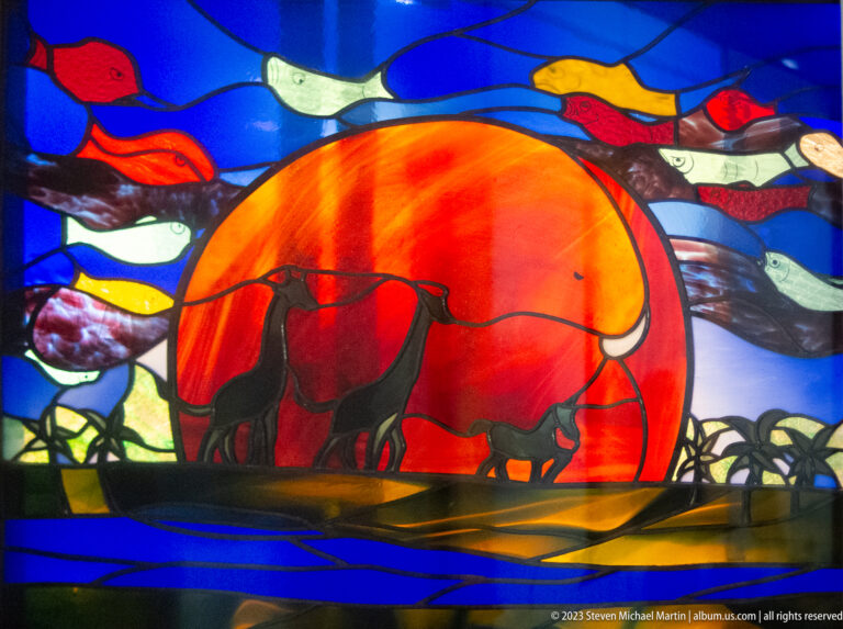 Stained-glass panel showing cattle before a large orange sun with colorful bands above.