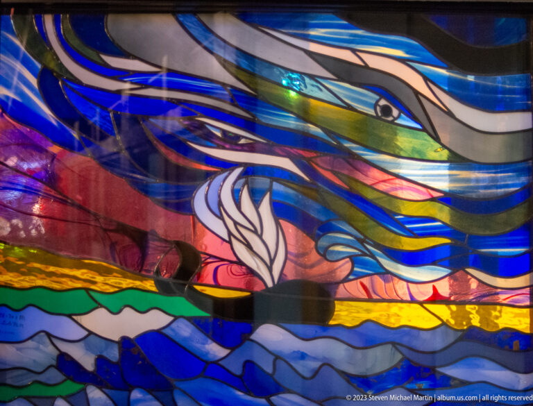 Colorful stained-glass panel with sweeping blue, white, pink, green, and gold forms.