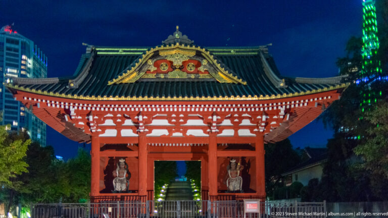 Illuminated red Sangedatsumon gate with guardian figures and part of Tokyo Tower at right.