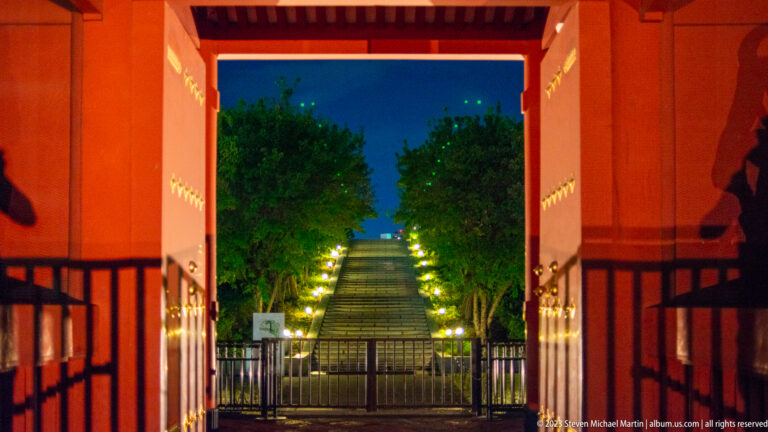 Illuminated stairway rising through trees, framed by a red temple gateway.