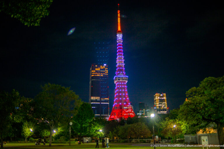 Tokyo Tower glowing red, purple, and orange beyond trees and a dark park lawn.