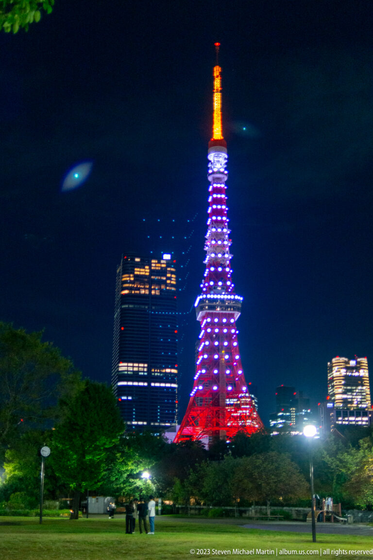 Closer park view of Tokyo Tower glowing red, violet, and orange above trees.