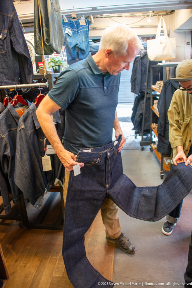 Man holds dark jeans against his waist inside a crowded clothing shop.