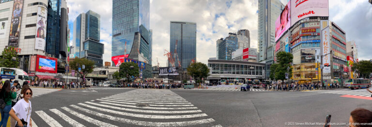 Wide view across Shibuya Crossing with empty lanes in front and crowds waiting beyond.