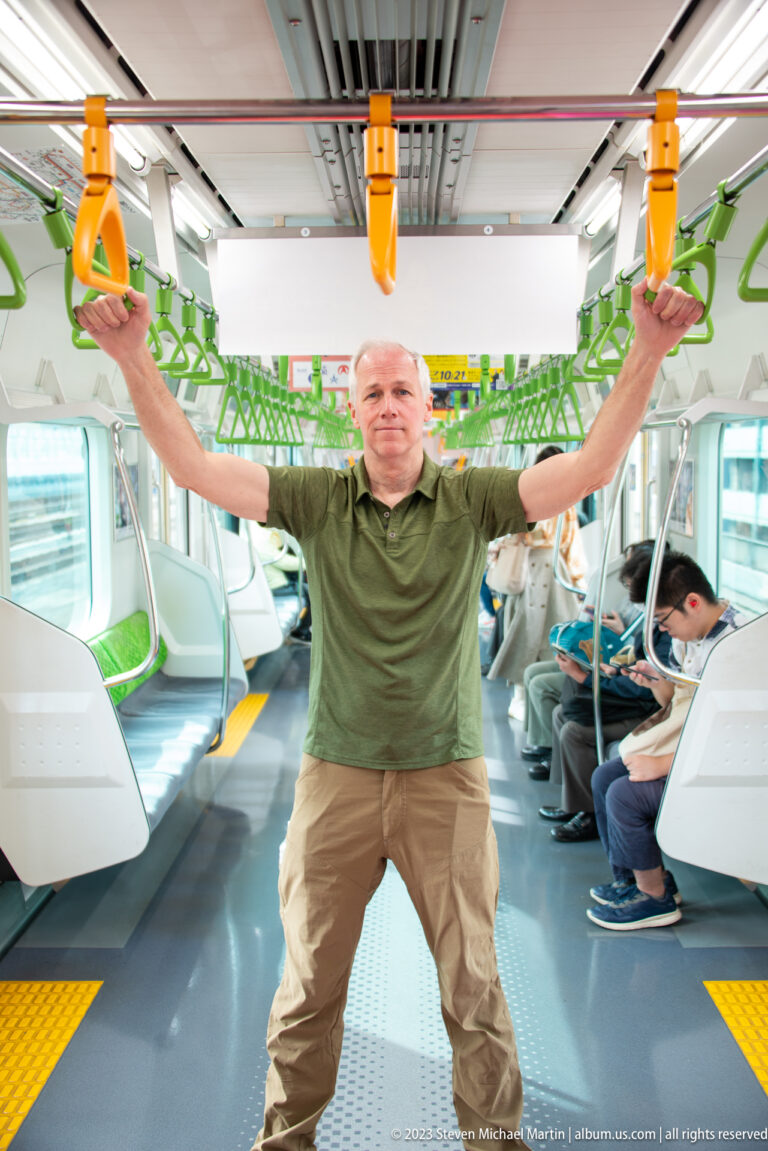 Man stands in a Tokyo train holding two overhead straps between rows of seats.
