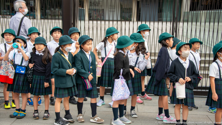 Uniformed Japanese schoolchildren in green hats stand together with bags and water bottles.