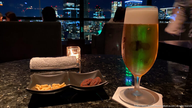 Tall beer glass, two snack dishes, candle, and towel on a lounge table with city lights behind.