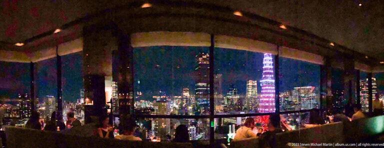 Tokyo Tower seen through lounge windows with reflections, seated guests, and city lights.