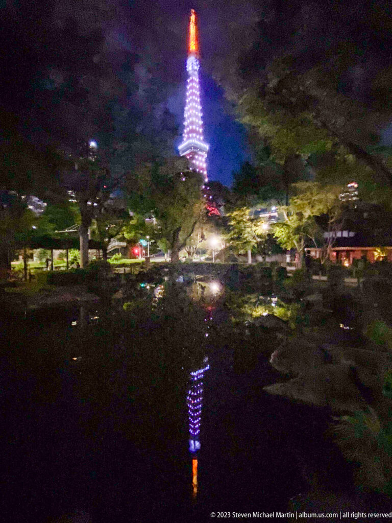 Tokyo Tower rises beyond trees with its colored reflection visible in a dark pond.