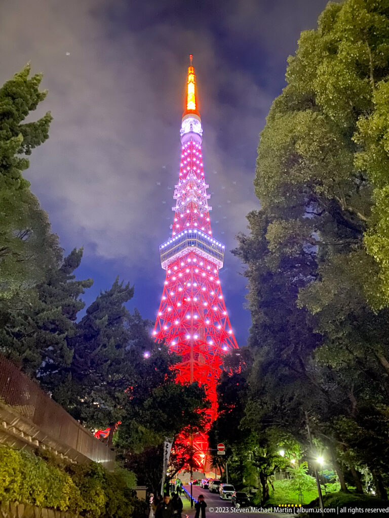 Tokyo Tower glows red and white between large trees along a landscaped night path.