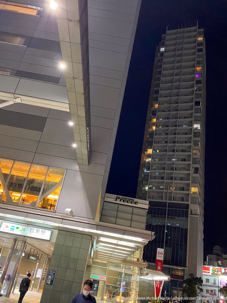 Night view of a tall modern tower above a brightly lit station entrance and canopy.