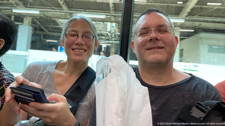 Two smiling travelers stand in an airport terminal holding a phone and shopping bag.